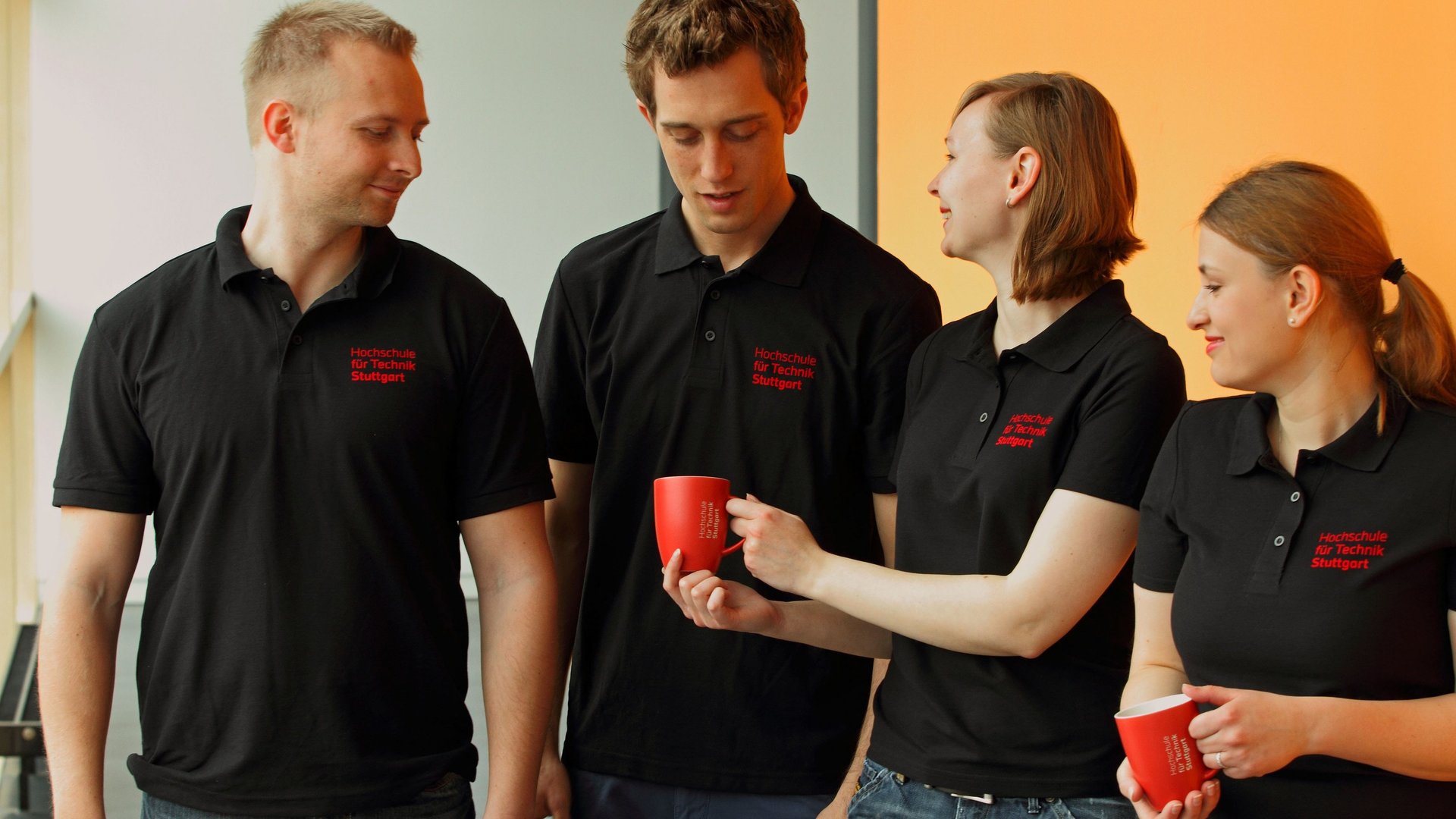 Vier Studierende tragen T-Shirts mit HFT Logo und halten HFT-Tassen in der Hand