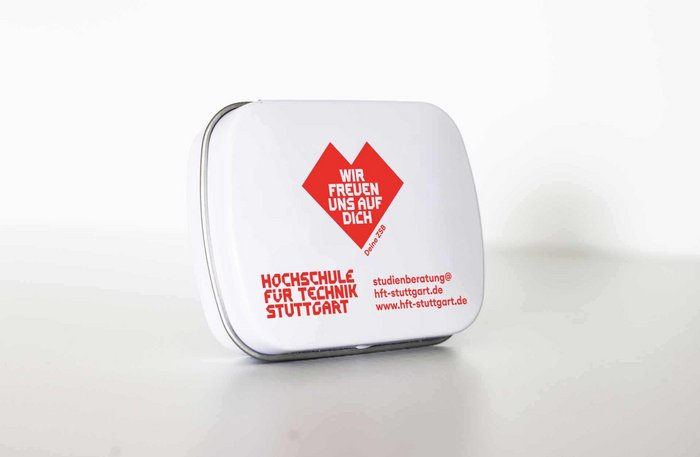 Weiße Bonbon-Dose aus Blech mit runden Ecken und einer roten Aufschrift "Wir freuen uns auf Dich" in einem roten Herz sowie das Logo der HFT Stuttgart