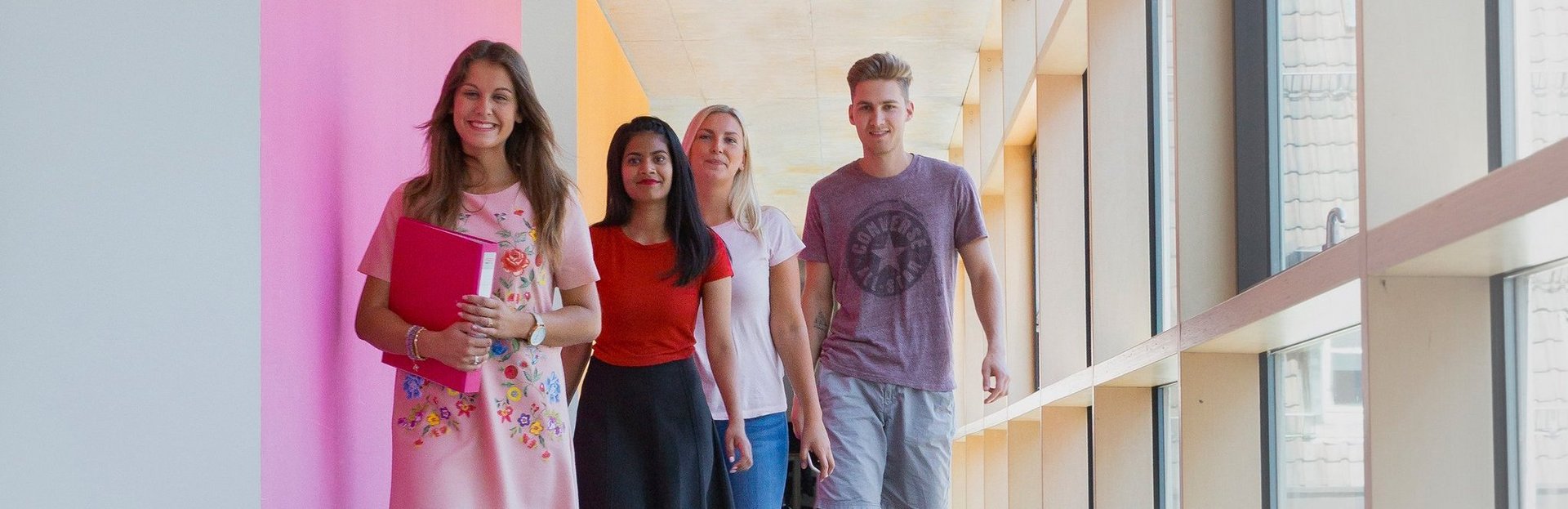 Drei Studierende laufen auf dem Gang in einem Gebäude der HFT Stuttgart. In ihren Armen halten Sie Bücher.