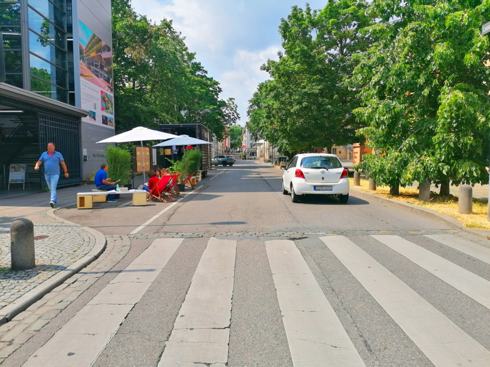 Blick vom Zebrastreifen auf eine baumbestandene Straße mit Aktionsständen auf der linken Seite 