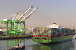 Einfahrt eines Containerschiffs in einen Hafen. Im Hintergrund stehen Kräne